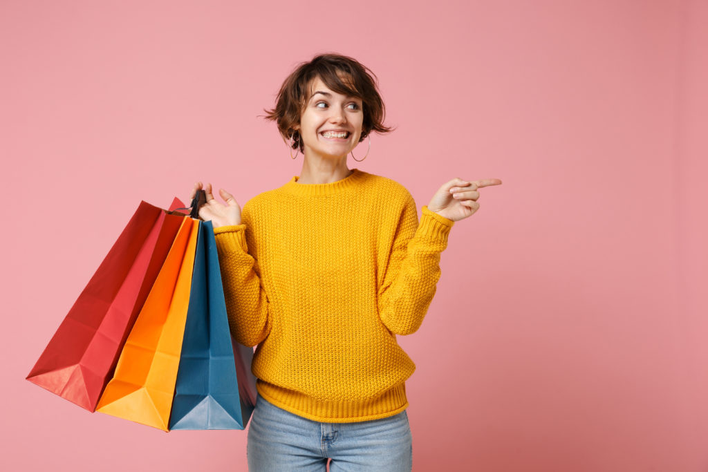 Smiling young buyer woman girl in yellow sweater posing isolated on pink background. People lifestyle concept. Mock up copy space. Hold package bag with purchases after shopping point finger aside