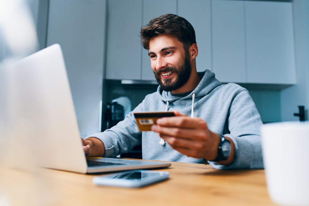 Cheerful young buyer man paying bills online with credit card and laptop