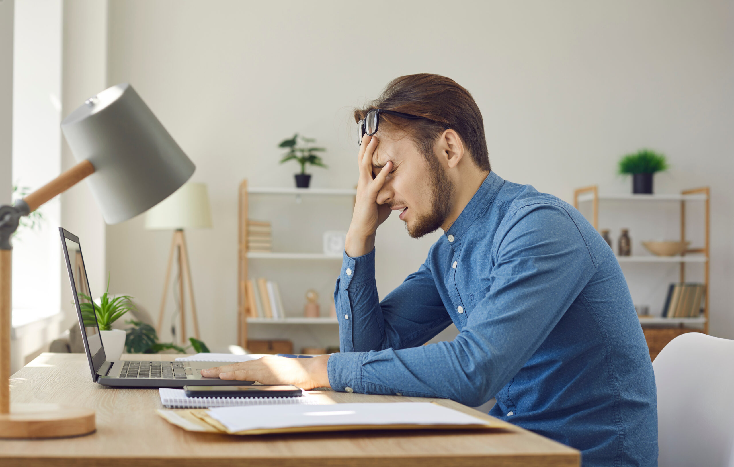 Tired worker having problem. Man makes dumb mistake or has difficulty with digital file. Side view of upset stressed young guy facepalming sitting at working desk with laptop computer in home office